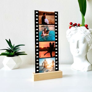 May include: A clear acrylic photo frame with a wooden base. The frame features a film strip design and holds multiple photos of a couple enjoying various activities, including a beach scene, a sunset, and a swimming pool.