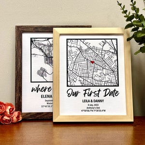 May include: Two framed map prints. One has a dark brown frame and the other has a light wood frame. The maps are black and white, with a red heart marking a location on one. Text on the prints reads "where" and "Our First Date."