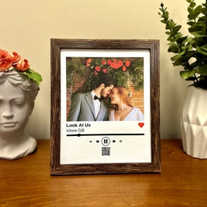 May include: A framed print of a couple in a wedding photo with a music player interface below the photo. The music player interface shows the song "Look At Us" by Vince Gill. The music player has a red heart icon, play and pause buttons, and a QR code.