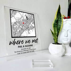 Puede incluir: Un letrero de acr&iacute;lico transparente con un mapa en blanco y negro de una ciudad. El mapa tiene un coraz&oacute;n rojo que marca una ubicaci&oacute;n. El texto "where we met" est&aacute; escrito en cursiva negra. Debajo del mapa, el texto "Phil & Suzanne" est&aacute; escrito en negro. El texto "November 24, 2001" est&aacute; escrito en negro. El texto "The University of Michigan Central Campus" est&aacute; escrito en negro. El texto "N 42&deg; 16' 37.681, W 83&deg; 44' 17.692" est&aacute; escrito en negro.