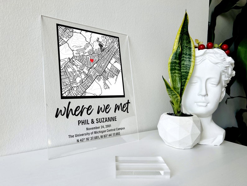 Puede incluir: Un letrero de acr&iacute;lico transparente con un mapa en blanco y negro de una ciudad. El mapa tiene un coraz&oacute;n rojo que marca una ubicaci&oacute;n. El texto "where we met" est&aacute; escrito en cursiva negra. Debajo del mapa, el texto "Phil & Suzanne" est&aacute; escrito en negro. El texto "November 24, 2001" est&aacute; escrito en negro. El texto "The University of Michigan Central Campus" est&aacute; escrito en negro. El texto "N 42&deg; 16' 37.681, W 83&deg; 44' 17.692" est&aacute; escrito en negro.