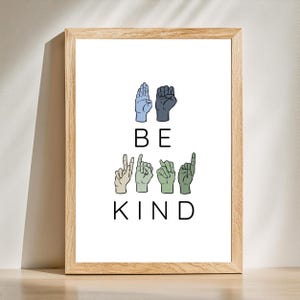 May include: A framed print with the words "BE KIND" in black text. The word "BE" is represented by hand gestures in blue and black. The word "KIND" is represented by hand gestures in various shades of green and tan. The frame is made of light brown wood.
