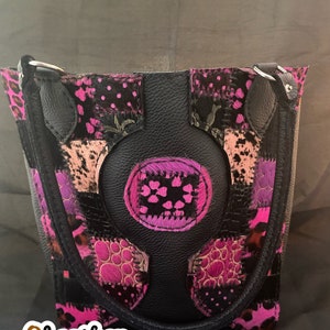 Op de afbeelding: Een zwarte leren tote bag met een patchwork ontwerp met roze, paarse en zwarte dierenprint stof. De tas heeft een groot rond detail in het midden en twee bovenste handvatten.