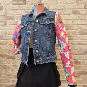 May include: A blue denim jacket with patchwork sleeves. The jacket has a button-up front and collar. The sleeves are made of patchwork fabric with pink, red, yellow, and floral patterns. The jacket is paired with a black skirt. The jacket is displayed on a mannequin.