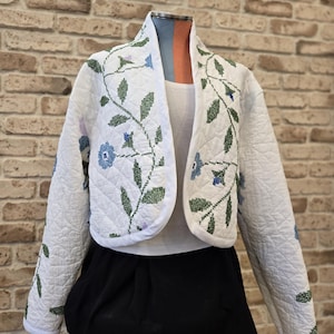 May include: White quilted jacket with a floral embroidery design featuring blue and green flowers and leaves. The jacket has a shawl collar and long sleeves.