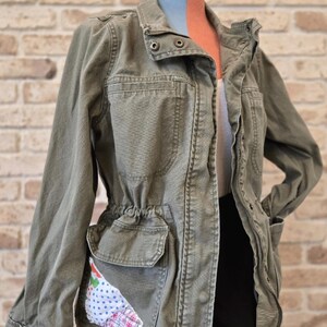 Könnte beinhalten: Olivgrüne Utility-Jacke mit Kordelzug in der Taille und mehreren Taschen. Die Jacke hat einen dekorativen Aufnäher auf einer Tasche mit Blumen- und Karomustern. Die Jacke wird auf einer Schaufensterpuppe vor einer Backsteinmauer präsentiert.
