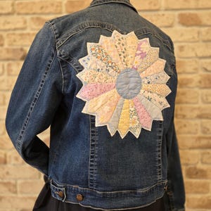 Può includere: Giacca di jeans blu scuro con un grande motivo floreale patchwork colorato sul retro. Il fiore presenta tessuti pastello nei toni del rosa, giallo, blu e bianco, creando un look unico e artigianale.