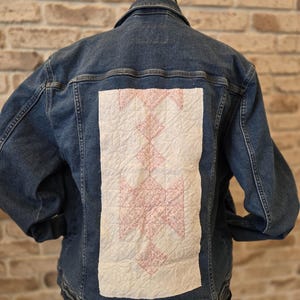 Può includere: Giacca di jeans blu con un design trapuntato bianco e rosa sul retro. La toppa trapuntata presenta un motivo geometrico nelle tonalità del rosa e del bianco. La giacca ha un colletto classico, chiusure con bottoni e tasche.