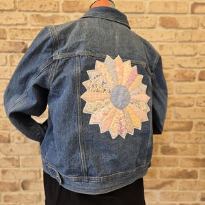 Può includere: Giacca di jeans blu con un grande motivo floreale trapuntato e colorato sul retro. Il fiore presenta tessuti pastello nelle tonalità del rosa, giallo e blu. La giacca è esposta su un manichino.