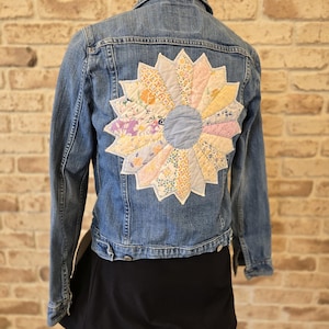 Può includere: Giacca di jeans blu chiaro con un grande, colorato, motivo a girasole trapuntato sul retro. Il girasole è realizzato con vari pezzi di tessuto fantasia dai colori pastello. La giacca ha un colletto classico e chiusure con bottoni.