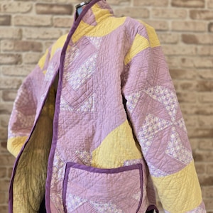 Puede incluir: Una chaqueta acolchada en tonos lavanda y amarillo, con ribete morado. La chaqueta presenta un diseño de patchwork con un bolsillo en la parte delantera. La chaqueta se exhibe en un maniquí contra una pared de ladrillos.