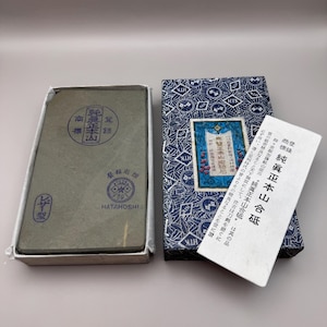 May include: A gray sharpening stone in a white box, next to a blue patterned box and a white paper with Japanese text. The stone has Japanese text and stamps. The blue box has a blue and white design and a label with Japanese text.