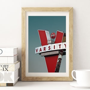 May include: A framed photograph of a red and white sign with the words "The Varsity" in white letters. The sign is shaped like a "V" and has a Coca-Cola logo on the top.