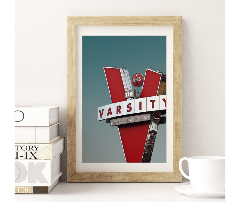 May include: A framed photograph of a red and white sign with the words "The Varsity" in white letters. The sign is shaped like a "V" and has a Coca-Cola logo on the top.