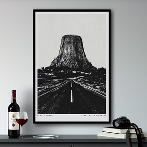 Könnte beinhalten: Schwarzweiß-Illustration des Devils Tower National Monument in den Black Hills, Wyoming. Das Bild zeigt eine Straße, die zur Turmfelsen führt, mit dem Text "Devils Tower" und "Black Hills, Wyoming" unten.