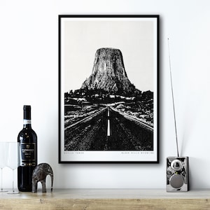 Könnte beinhalten: Schwarzweißfoto des Devils Tower National Monument in den Black Hills, Wyoming. Das Bild zeigt eine Straße, die zur Spitze führt, mit Bäumen und Büschen im Vordergrund.