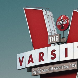May include: A red and white sign with the words "THE VARSITY" in large white letters. The sign also features a red Coca-Cola logo and the text "Coca-Cola Salutes THE VARSITY Since 1928".
