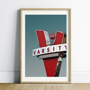 May include: A red and white sign with the words "The Varsity" and "Serving the Varsity Since 1928" in white letters. The sign is shaped like a "V" and has a Coca-Cola logo on top.