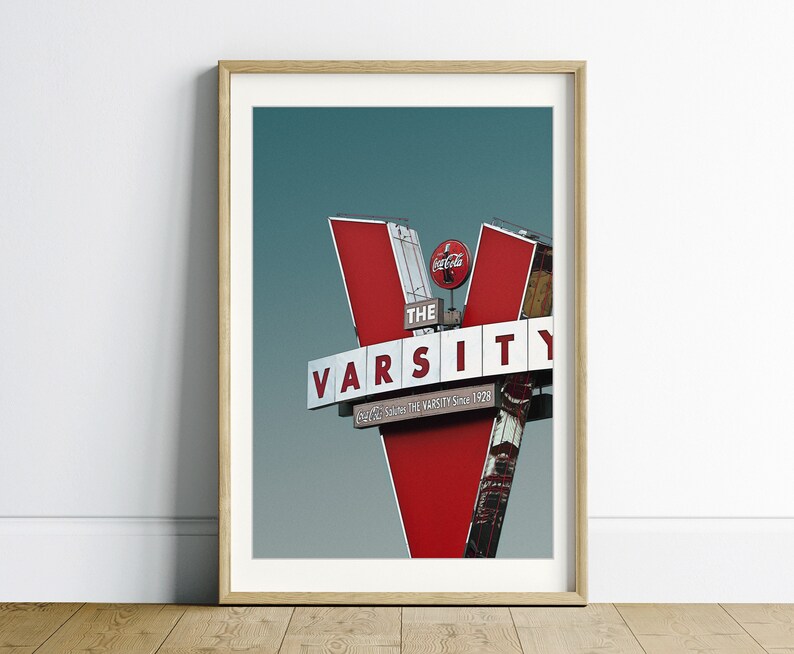 May include: A red and white sign with the words "The Varsity" and "Serving the Varsity Since 1928" in white letters. The sign is shaped like a "V" and has a Coca-Cola logo on top.