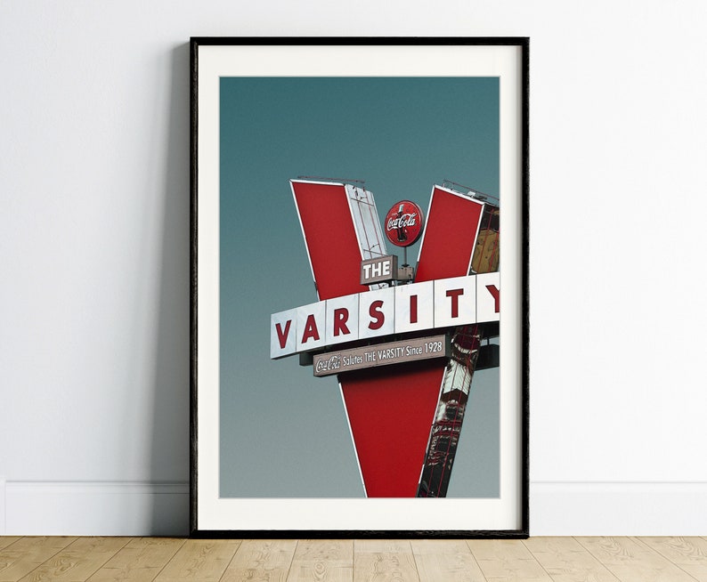 May include: A red and white sign with the words "The Varsity" in white letters. The sign is shaped like a V and has a Coca-Cola logo on the top. The sign is against a blue sky.