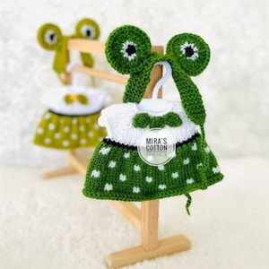 May include: Handmade green and white crochet dress with white polka dots, displayed on a wooden hanger. The dress features a white top and a green skirt. The hanger is attached to a wooden stand. The text "MIRA'S COTTON FRIENDS" is visible.