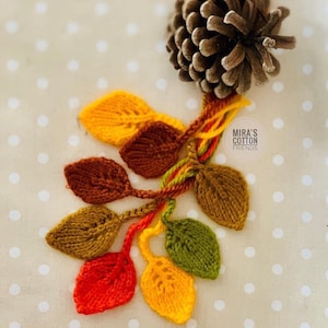 May include: A knitted leaf garland in autumnal colours, with a pine cone. The leaves are in shades of red, yellow, brown, and green. The text "MIRA'S COTTON FRIENDS" is visible.