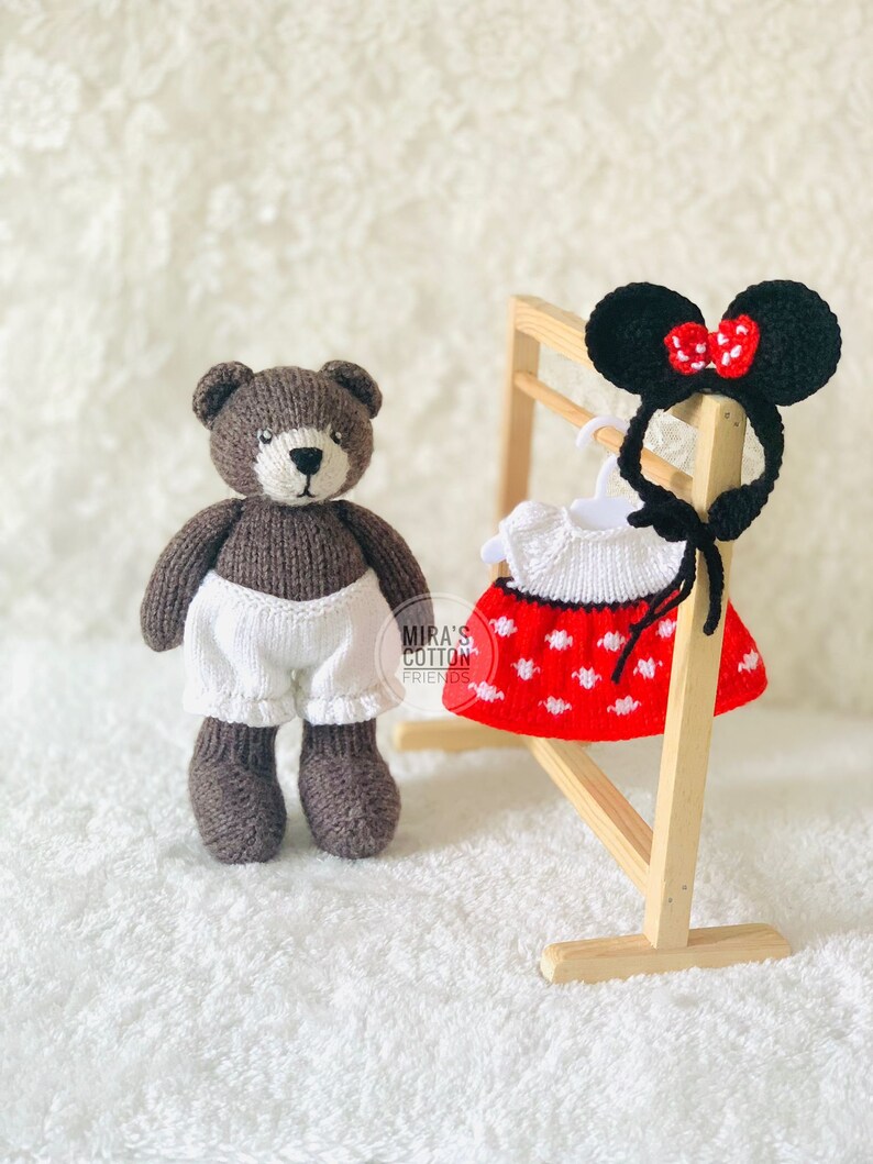 K&ouml;nnte beinhalten: Ein brauner, gestrickter Teddyb&auml;r mit wei&szlig;en Shorts steht neben einem Holzst&auml;nder mit einem rot-wei&szlig; gepunkteten Kleid und schwarzen M&auml;useohren mit roter Schleife. Der Text "Mira's Cotton Friends" ist sichtbar.