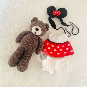 K&ouml;nnte beinhalten: Ein gestrickter brauner Teddyb&auml;r mit wei&szlig;er Schnauze und passendem Outfit. Das Outfit umfasst ein rotes Kleid mit wei&szlig;en Punkten, wei&szlig;e Pumphosen und ein schwarzes Stirnband mit roter Schleife. Die Artikel liegen auf einer wei&szlig;en Oberfl&auml;che.