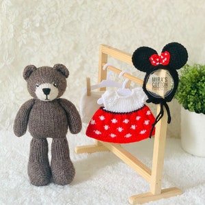 K&ouml;nnte beinhalten: Ein brauner, gestrickter Teddyb&auml;r steht neben einer Holzgarderobe mit einem wei&szlig;en Kleid mit roten Punkten, einer wei&szlig;en Shorts und einem schwarzen Minnie-Mouse-Ohren-Haarreif mit roter Schleife. Der Haarreif tr&auml;gt den Text "Mira's Cotton Friends".