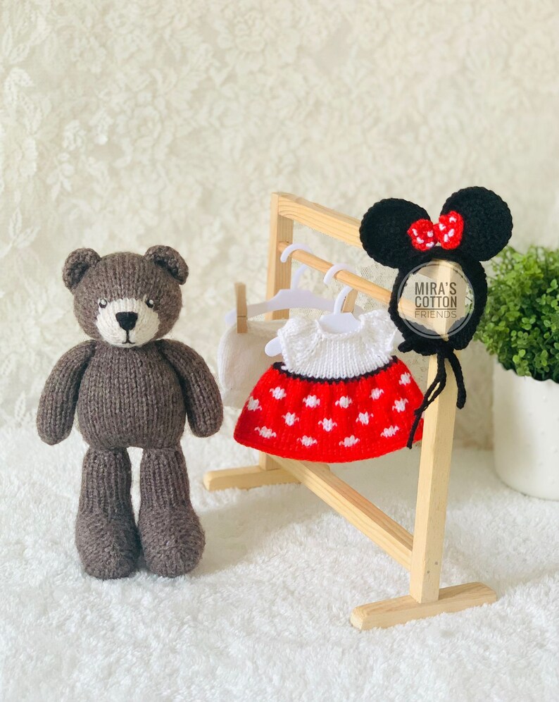 K&ouml;nnte beinhalten: Ein brauner, gestrickter Teddyb&auml;r steht neben einer Holzgarderobe mit einem wei&szlig;en Kleid mit roten Punkten, einer wei&szlig;en Shorts und einem schwarzen Minnie-Mouse-Ohren-Haarreif mit roter Schleife. Der Haarreif tr&auml;gt den Text "Mira's Cotton Friends".