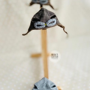 May include: Hand-knitted aviator hats in brown and gray, each featuring goggles. The hats are displayed on wooden stands against a soft, dotted background. The hats have ear flaps and are designed for babies.