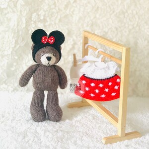 K&ouml;nnte beinhalten: Ein handgefertigter brauner Teddyb&auml;r mit schwarzem Hut und roter Schleife. Ein rot-wei&szlig; gepunktetes Kleid h&auml;ngt an einem Holzgestell. Das Kleid hat einen wei&szlig;en Kragen. Der Hintergrund ist eine wei&szlig;e, strukturierte Oberfl&auml;che.