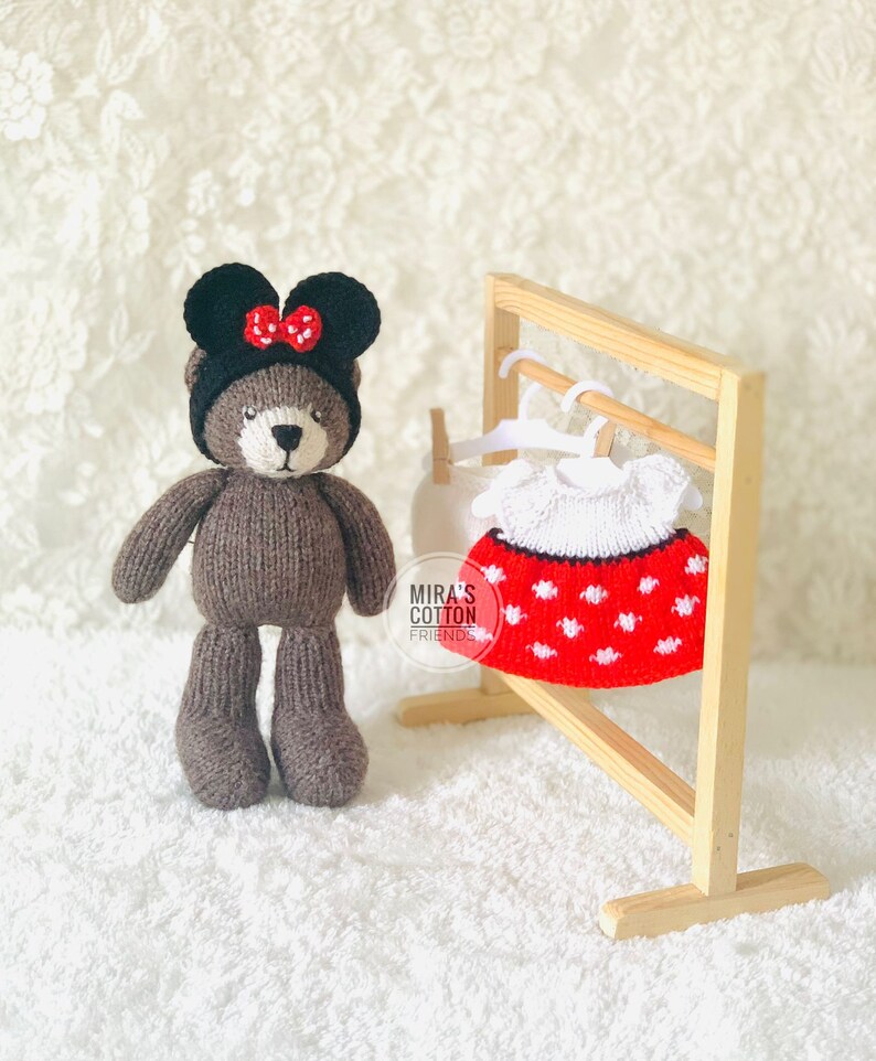 K&ouml;nnte beinhalten: Ein handgefertigter brauner Teddyb&auml;r mit schwarzem Hut und roter Schleife. Ein rot-wei&szlig; gepunktetes Kleid h&auml;ngt an einem Holzgestell. Das Kleid hat einen wei&szlig;en Kragen. Der Hintergrund ist eine wei&szlig;e, strukturierte Oberfl&auml;che.