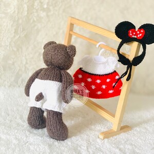 K&ouml;nnte beinhalten: Ein brauner, gestrickter Teddyb&auml;r mit wei&szlig;en Shorts steht neben einem Holzgestell mit einem rot-wei&szlig; gepunkteten Kleid und einem schwarzen Stirnband mit M&auml;useohren und einer roten Schleife. Der Text "MIRA'S COTTON FRIENDS" ist sichtbar.