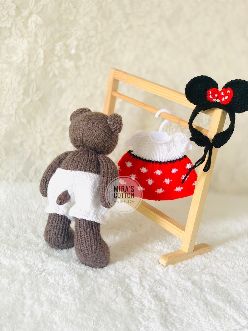 K&ouml;nnte beinhalten: Ein brauner, gestrickter Teddyb&auml;r mit wei&szlig;en Shorts steht neben einem Holzgestell mit einem rot-wei&szlig; gepunkteten Kleid und einem schwarzen Stirnband mit M&auml;useohren und einer roten Schleife. Der Text "MIRA'S COTTON FRIENDS" ist sichtbar.