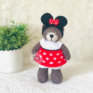 K&ouml;nnte beinhalten: Ein handgefertigter brauner Teddyb&auml;r mit einem schwarzen Stirnband mit M&auml;useohren und einer roten Schleife. Der B&auml;r tr&auml;gt ein rotes Kleid mit wei&szlig;en Punkten und einen wei&szlig;en Kragen. Der Text "Mira's Cotton Friends" ist sichtbar.