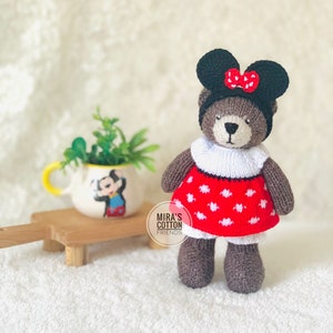 K&ouml;nnte beinhalten: Ein handgefertigter brauner Teddyb&auml;r mit schwarzem Hut und roter Schleife, wei&szlig;em Kragen und rotem Kleid mit wei&szlig;en Punkten. Der B&auml;r steht auf einer wei&szlig;en Oberfl&auml;che. Im Hintergrund ein kleiner Keramiktopf mit Mickey Mouse-Design.