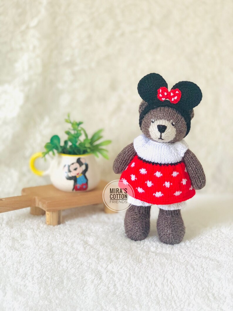 K&ouml;nnte beinhalten: Ein handgefertigter brauner Teddyb&auml;r mit schwarzem Hut und roter Schleife, wei&szlig;em Kragen und rotem Kleid mit wei&szlig;en Punkten. Der B&auml;r steht auf einer wei&szlig;en Oberfl&auml;che. Im Hintergrund ein kleiner Keramiktopf mit Mickey Mouse-Design.
