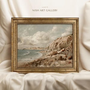 May include: A framed landscape painting of a coastal scene. The artwork depicts a beach with a rocky cliffside, a town in the distance, and a cloudy sky. The frame is gold-colored, and the text "LUCY NISH ART GALLERY" is visible.
