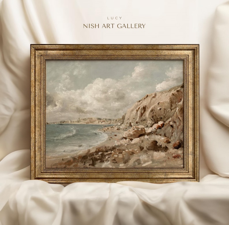 May include: A framed landscape painting of a coastal scene. The artwork depicts a beach with a rocky cliffside, a town in the distance, and a cloudy sky. The frame is gold-colored, and the text "LUCY NISH ART GALLERY" is visible.