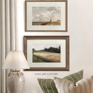 May include: Two framed landscape paintings with gold-toned frames hang on a white wall. The top painting depicts a seascape with a cloudy sky. The bottom painting shows a field of wheat. A lamp and pillows are in the foreground. The text "NISH ART GALLERY" is visible.
