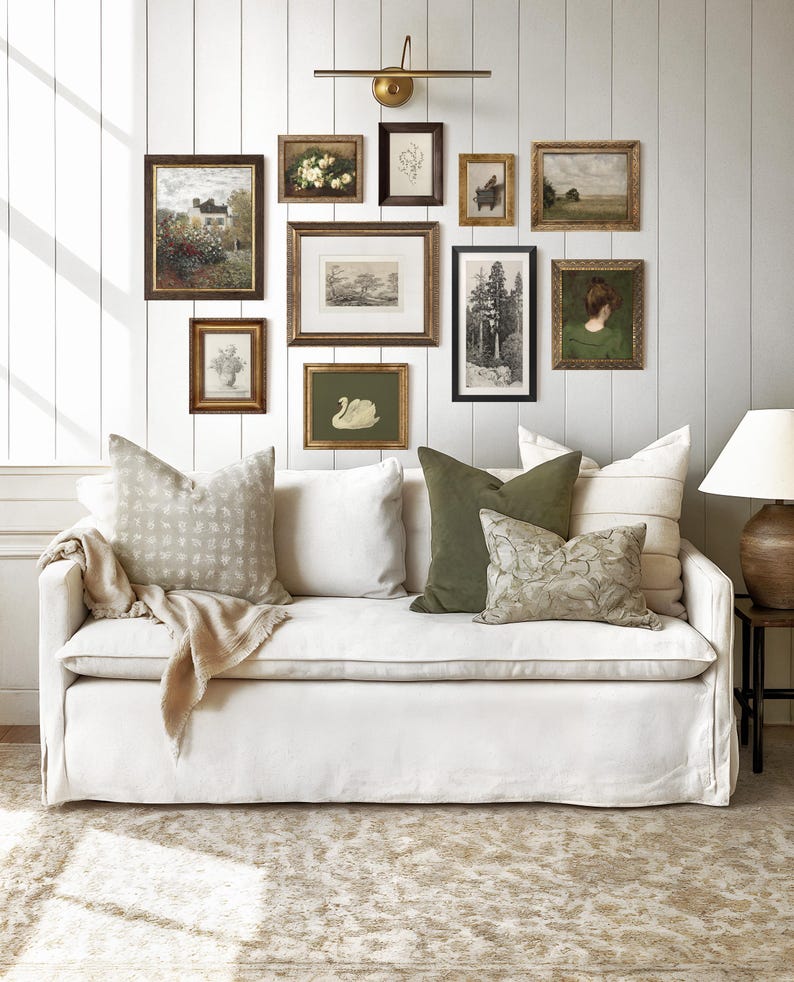 May include: A living room scene featuring a white sofa adorned with decorative pillows and a collection of framed artwork on the wall. The artwork includes landscape paintings, botanical prints, and a swan illustration. A brass wall lamp illuminates the art.