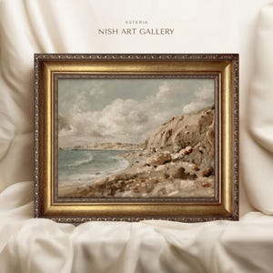 May include: A framed landscape painting of a coastal scene. The artwork features a beach with waves, cliffs, and a town in the distance. The frame is ornate with a gold finish. The text "ASTERIA NISH ART GALLERY" is at the top.
