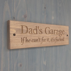 May include: A rectangular wooden sign with the words "Dad's Garage" and the phrase "If he can't fix it, it's fucked" engraved in a serif font. The sign is mounted on a blue-gray wooden surface.
