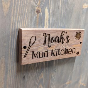 May include: Wooden sign with the text "Noah's Mud Kitchen" in a rustic font. The sign is brown and has a spoon and a paint splatter design.