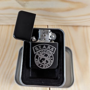 Puede incluir: Un encendedor negro con un dise&ntilde;o grabado en plata del logotipo del Departamento de Polic&iacute;a de Raccoon City S.T.A.R.S. El encendedor est&aacute; en una caja de regalo forrada de terciopelo negro.