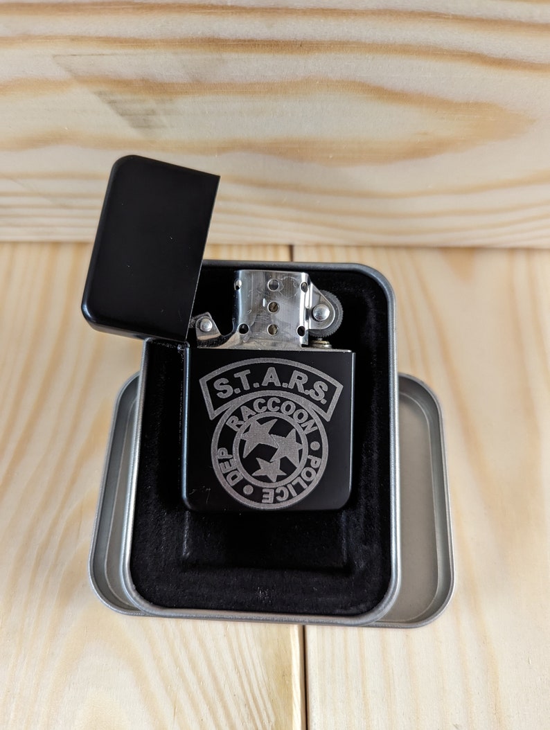 Puede incluir: Un encendedor negro con un dise&ntilde;o grabado en plata del logotipo del Departamento de Polic&iacute;a de Raccoon City S.T.A.R.S. El encendedor est&aacute; en una caja de regalo forrada de terciopelo negro.