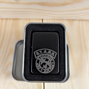 Puede incluir: Un encendedor negro con un dise&ntilde;o grabado en plata del logotipo de S.T.A.R.S. de la serie de videojuegos Resident Evil. El logotipo presenta las palabras "S.T.A.R.S." y "Raccoon Police Department" con una estrella y dos alas.