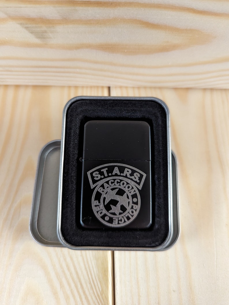Puede incluir: Un encendedor negro con un dise&ntilde;o grabado en plata del logotipo de S.T.A.R.S. de la serie de videojuegos Resident Evil. El logotipo presenta las palabras "S.T.A.R.S." y "Raccoon Police Department" con una estrella y dos alas.
