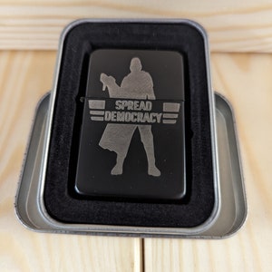 May include: A black lighter with a silver design of a person holding a weapon and the text "SPREAD DEMOCRACY". The lighter is in a silver tin.